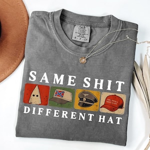 Könnte beinhalten: Ein graues Comfort Colors T-Shirt mit dem weißen Text "SAME SHIT DIFFERENT HAT". Das Shirt zeigt vier Bilder von Hüten, darunter eine KKK-Haube und einen "Make America Great Again"-Hut. Eine silberne Halskette ist über dem Shirt drapiert.