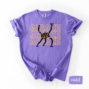 May include: Violet-colored t-shirt with the word "AMAZE" stacked three times in a yellow font. A black, stylized figure with outstretched arms and legs is centered on the shirt. The shirt has rolled-up sleeves.