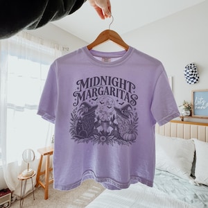 May include: A light purple short-sleeve t-shirt featuring a dark graphic print with the text "MIDNIGHT MARGARITAS" above two figures in witch hats, a cat, a pumpkin, and a full moon with bats, all surrounded by botanical elements.
