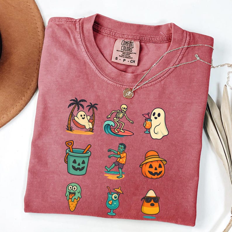 May include: A maroon Comfort Colors t-shirt featuring a variety of Halloween-themed illustrations. The graphics include a ghost relaxing in a hammock, a skeleton surfing, and a jack-o'-lantern. A silver necklace with a pendant is draped across the shirt.