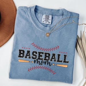 May include: A light blue t-shirt with a distressed look featuring a black graphic of two baseball bats and the text "Baseball mom" with red stitching around the text and bats.