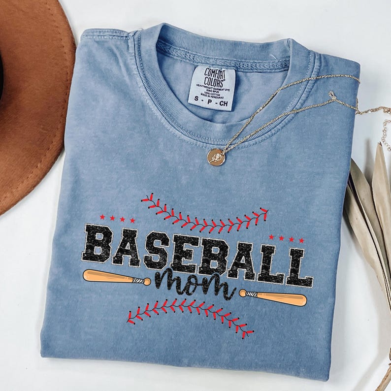 May include: A light blue t-shirt with a distressed look featuring a black graphic of two baseball bats and the text "Baseball mom" with red stitching around the text and bats.