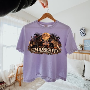 May include: A light purple t-shirt featuring a graphic print of two witches in hats sitting by a lake under a full moon, holding margaritas. The design includes a black cat, a broom, candles, a skull, and the text Midnight Margaritas in a script font.