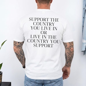 Può includere: T-shirt bianca con la scritta "SUPPORT THE COUNTRY YOU LIVE IN OR LIVE IN THE COUNTRY YOU SUPPORT" stampata in nero. La maglietta è indossata da una persona con tatuaggi sulle braccia.