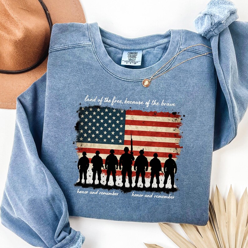 May include: A blue sweatshirt with a faded look features a graphic of an American flag with a line of soldiers in silhouette. The text "land of the free, because of the brave" is above the flag and "honor and remember" is below the flag.
