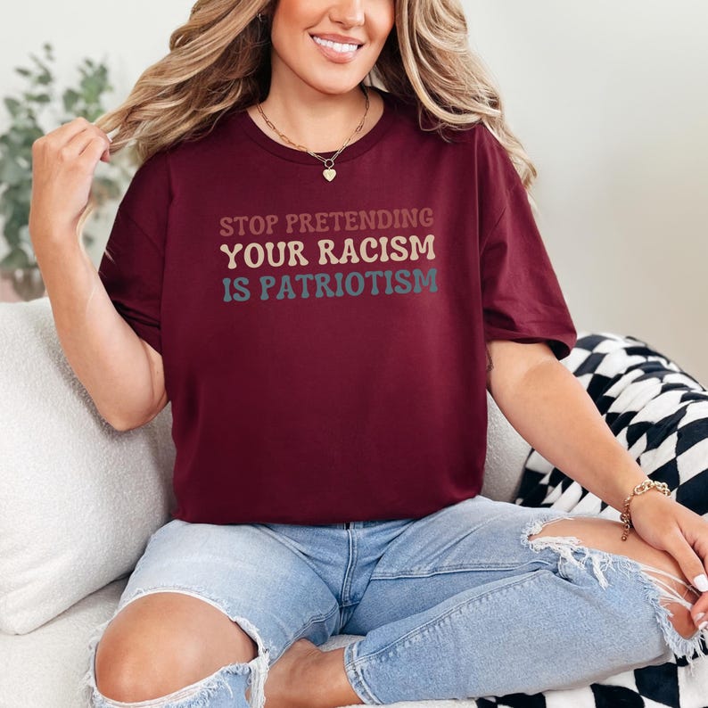 Pode incluir: Uma camiseta vinho com o texto "STOP PRETENDING YOUR RACISM IS PATRIOTISM" em uma fonte retr&ocirc; com letras de diferentes cores.