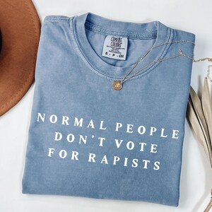 以下が含まれることがあります： 「NORMAL PEOPLE DON'T VOTE FOR RAPISTS」の白い文字が入った、青色のComfort Colors Tシャツ。ペンダント付きのゴールドネックレスがシャツの上にかけられています。茶色の帽子と乾燥した葉が背景にあります。