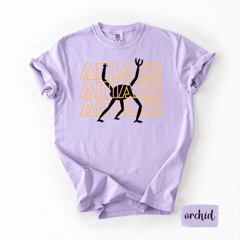May include: A light purple t-shirt with the word "AMAZE" repeated three times in a yellow-orange color. A black graphic of a stick figure is in the center. The shirt is a soft, comfortable material.