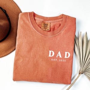 May include: A coral-colored t-shirt with the word "DAD" and the year "EST. 2025" printed in white. The shirt is folded and has a Comfort Colors tag. A brown hat and a dried palm leaf are in the background.
