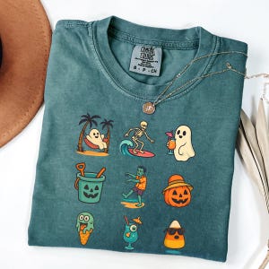May include: Teal Comfort Colors t-shirt with a variety of Halloween-themed graphics. The graphics include a ghost in a hammock, a skeleton surfing, a ghost with a drink, a jack-o'-lantern bucket, a zombie, a pumpkin with a hat, an ice cream cone, a cocktail, and a candy corn.