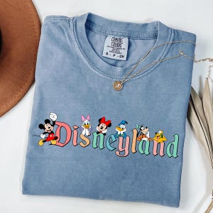 May include: A blue t-shirt featuring the word "Disneyland" in colorful letters with cartoon characters. The shirt has a crew neck and short sleeves. A gold necklace with a pendant is draped across the shirt.
