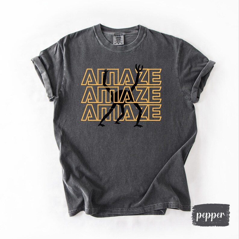 May include: Dark grey t-shirt with the word "AMAZE" in yellow, stacked three times, with a black silhouette figure in the center. The shirt has a crew neck and rolled sleeves. The word "pepper" is in the bottom right corner.