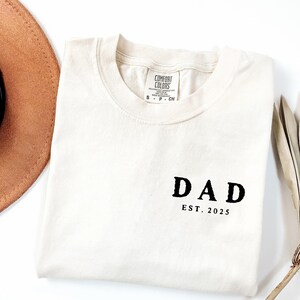 May include: Cream-colored t-shirt with the word "DAD" and "EST. 2025" printed in black. The shirt is folded and has a Comfort Colors tag. A brown hat and a feather are partially visible in the background.