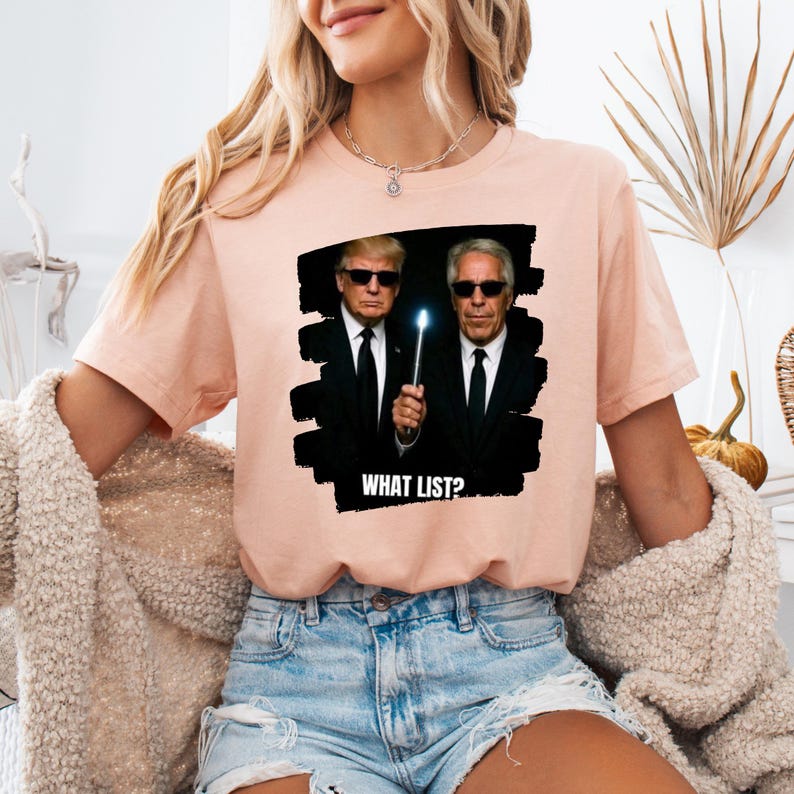 K&ouml;nnte beinhalten: Hellrosa T-Shirt mit einer Grafik von zwei M&auml;nnern in schwarzen Anz&uuml;gen und Sonnenbrillen, einer h&auml;lt einen leuchtenden Zauberstab. Der Text "WHAT LIST?" befindet sich unter dem Bild. Das T-Shirt wird von einer Person mit blonden Haaren und blauen Jeansshorts getragen.