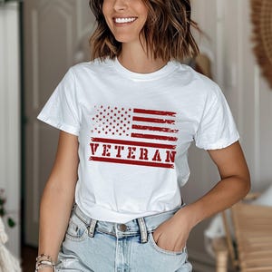 以下が含まれることがあります： 赤、白、青のくすんだアメリカ国旗のグラフィックと、赤い「VETERAN」の文字がプリントされた白いTシャツ