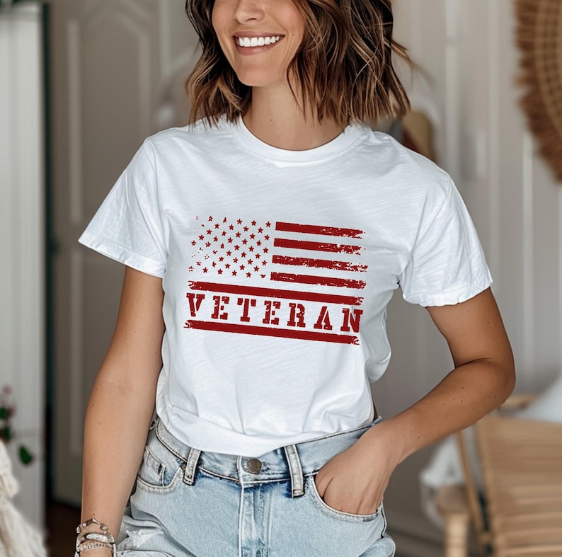 以下が含まれることがあります： 赤、白、青のくすんだアメリカ国旗のグラフィックと、赤い「VETERAN」の文字がプリントされた白いTシャツ