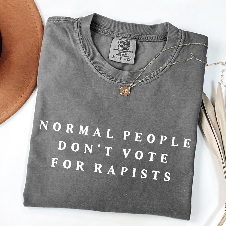 以下が含まれることがあります： 「NORMAL PEOPLE DON'T VOTE FOR RAPISTS」の白文字がプリントされたグレーのComfort Colors Tシャツ。小さなペンダントが付いたシルバーのネックレスがシャツの上にあります。茶色の帽子が左上に一部見えています。