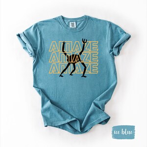 May include: Ice blue t-shirt with the word "AMAZE" in yellow, stacked three times, and a black stick figure graphic. The shirt has rolled-up sleeves and a crew neck.