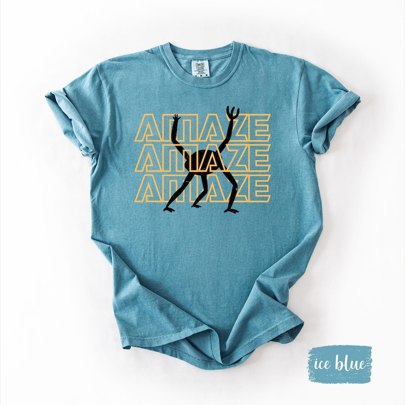 May include: Ice blue t-shirt with the word "AMAZE" in yellow, stacked three times, and a black stick figure graphic. The shirt has rolled-up sleeves and a crew neck.