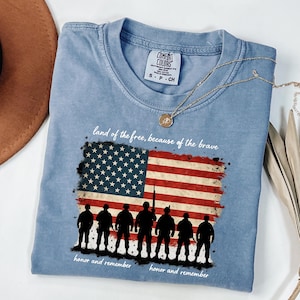 May include: A light blue t-shirt with a distressed American flag design and a silhouette of soldiers. The text "land of the free, because of the brave" and "honor and remember" is printed on the shirt.