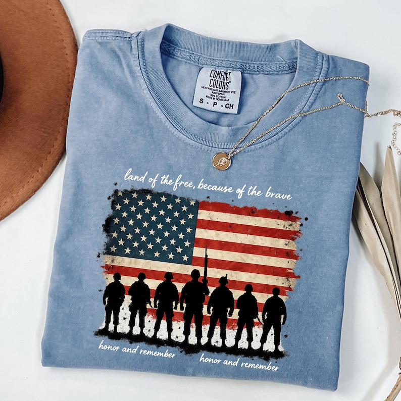 May include: A light blue t-shirt with a distressed American flag design and a silhouette of soldiers. The text "land of the free, because of the brave" and "honor and remember" is printed on the shirt.