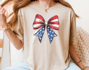 May include: A tan t-shirt featuring a patriotic bow design. The bow is rendered in red and white stripes and blue with white stars, evoking the American flag. The shirt is a casual, everyday wear item.