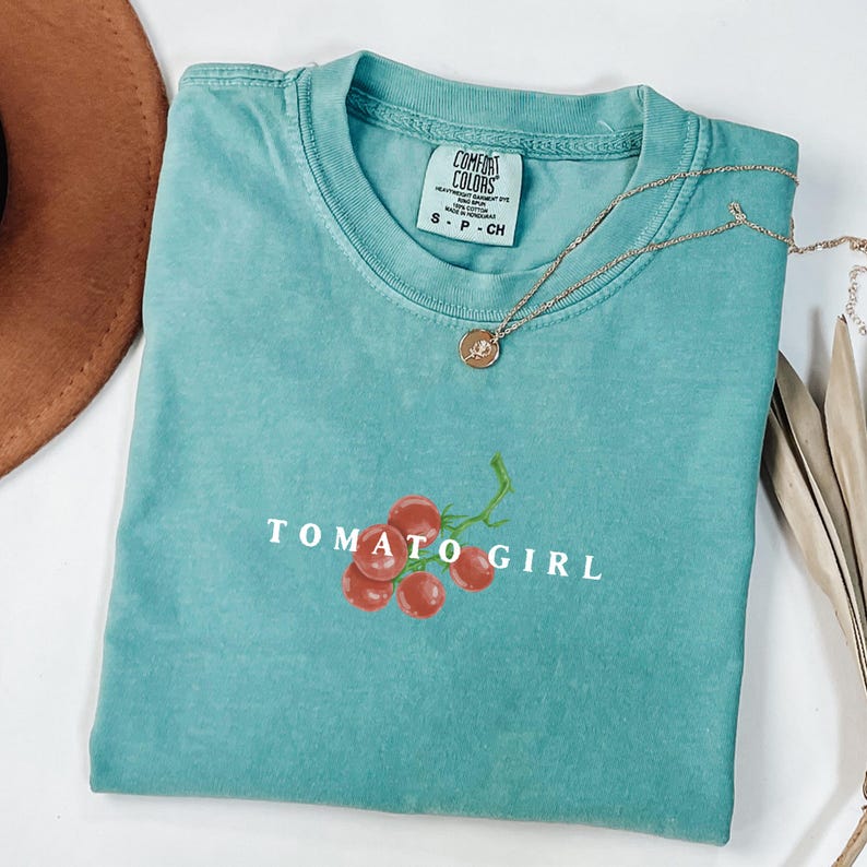 May include: A teal blue t-shirt with a white graphic of a tomato vine with red tomatoes and the text "TOMATO GIRL".