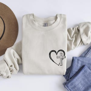 May include: A beige sweatshirt with a black heart and cross design. The heart is outlined and the cross is inside the heart.