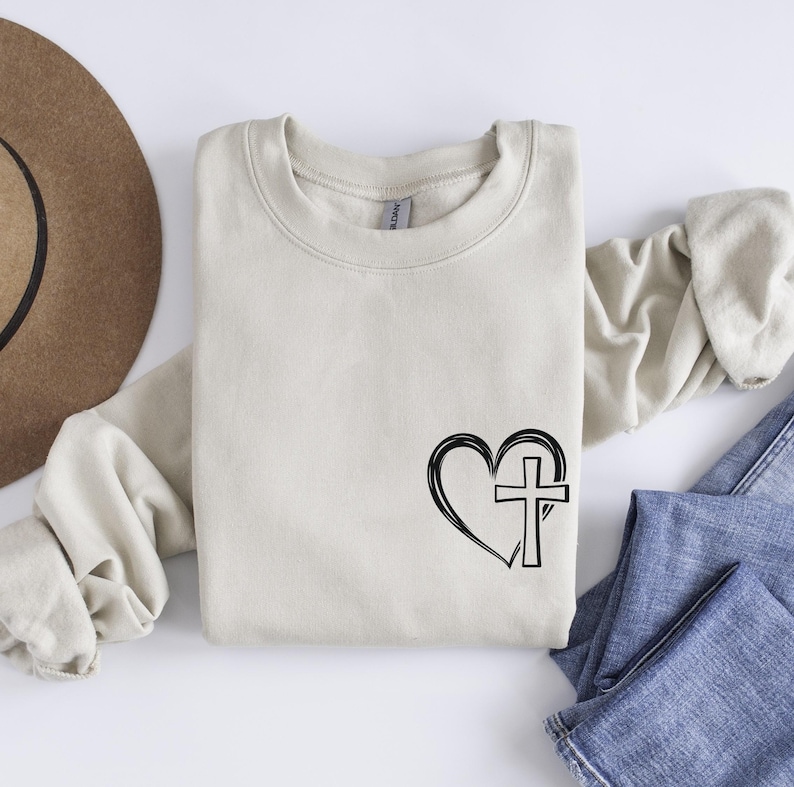May include: A beige sweatshirt with a black heart and cross design. The heart is outlined and the cross is inside the heart.