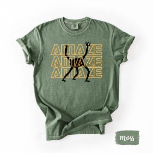 May include: Moss green t-shirt with the word "AMAZE" stacked three times in a yellow font, with a black stick figure design in the center. The shirt has rolled-up sleeves and a crew neck.
