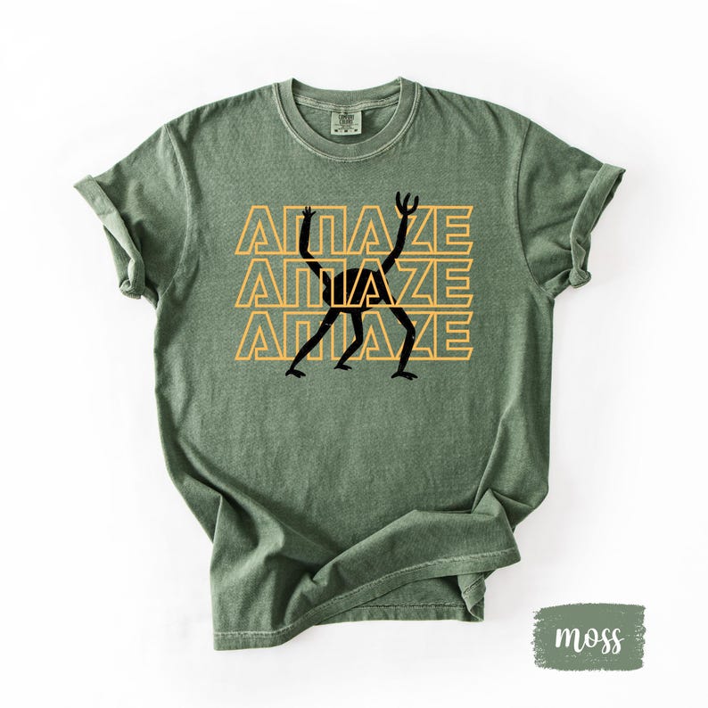 May include: Moss green t-shirt with the word "AMAZE" stacked three times in a yellow font, with a black stick figure design in the center. The shirt has rolled-up sleeves and a crew neck.