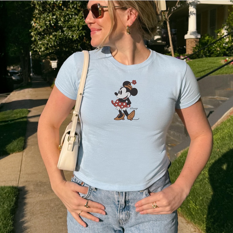 May include: Light blue t-shirt featuring a Minnie Mouse graphic. The cartoon character is depicted in a red and white polka dot dress, brown shoes, and a flower on her head. The shirt is paired with light wash denim jeans.