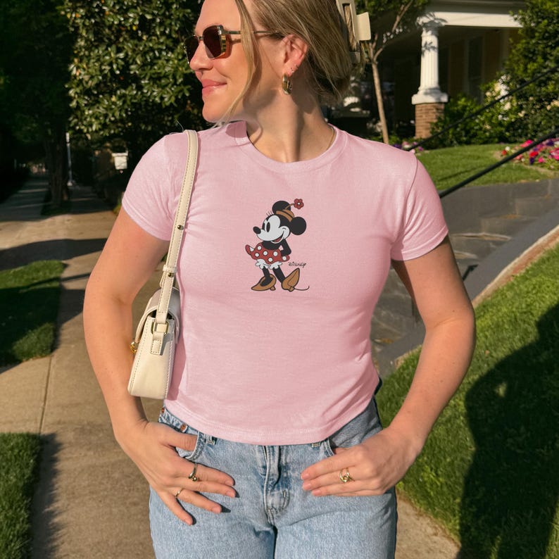 May include: A light pink t-shirt featuring a Minnie Mouse graphic. The graphic shows Minnie Mouse in a red polka dot dress and a matching bow. The shirt is paired with blue jeans.