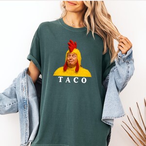 May include: Dark green t-shirt featuring a humorous graphic. The graphic depicts a person's face inside a yellow chicken costume with a red comb. The word "TACO" is printed in white below the image. The shirt has short sleeves.