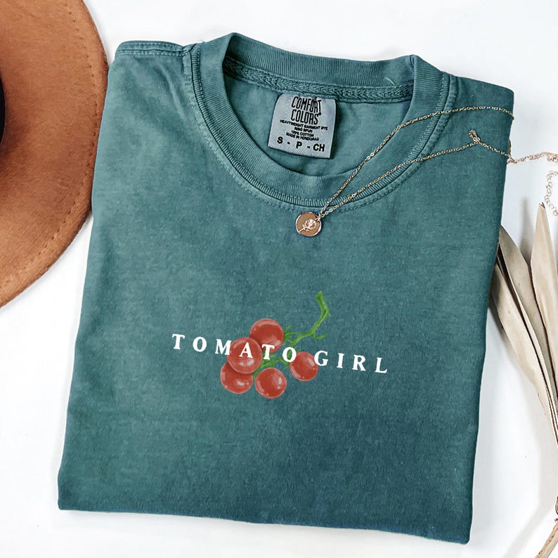 May include: A teal green t-shirt with a white graphic of a cluster of red tomatoes with a vine and the text "TOMATO GIRL".