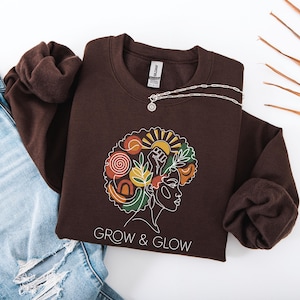 May include: A brown sweatshirt with a graphic of a woman's silhouette with a colorful afro, a sun, and the text "GROW & GLOW." A silver necklace with a pendant is draped across the sweatshirt. The sweatshirt is folded on top of a pair of blue jeans.