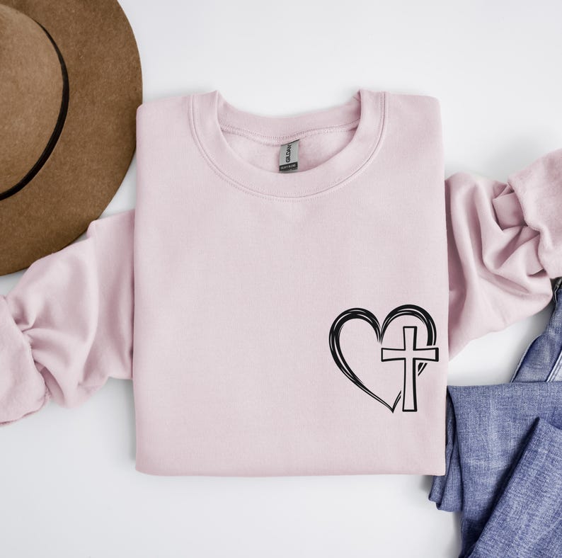 May include: A pink sweatshirt with a black heart and cross design. The heart is outlined and the cross is inside the heart.