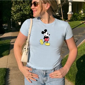May include: Light blue t-shirt featuring a classic Mickey Mouse graphic. The cartoon character is depicted in black, red, and yellow. The shirt has short sleeves and a crew neck. The person is wearing sunglasses and blue jeans.