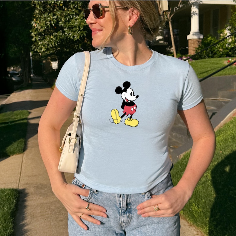 May include: Light blue t-shirt featuring a classic Mickey Mouse graphic. The cartoon character is depicted in black, red, and yellow. The shirt has short sleeves and a crew neck. The person is wearing sunglasses and blue jeans.