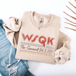 May include: A beige sweatshirt with the text "WSQK The Squawk 94.5 FM" in red and other colors. The sweatshirt is folded and placed on a white surface next to a pair of blue jeans.