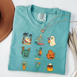 May include: Teal Comfort Colors t-shirt with a variety of Halloween-themed graphics. The graphics include a ghost in a hammock, a skeleton surfing, a ghost with a cocktail, a jack-o'-lantern bucket, a zombie, a pumpkin with a hat, ice cream, a cocktail, and candy corn.