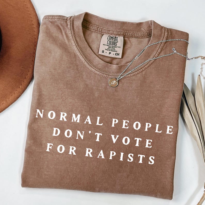 以下が含まれることがあります： 「NORMAL PEOPLE DON'T VOTE FOR RAPISTS」の白文字がプリントされた茶色のComfort Colors Tシャツ。小さなペンダントが付いたシルバーのネックレスがシャツの上にかけられています。左上には茶色の帽子が見えます。
