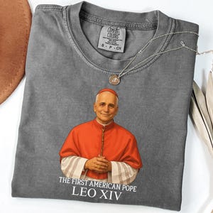 May include: A gray Comfort Colors t-shirt featuring a portrait of a man in a red robe, with the text "THE FIRST AMERICAN POPE LEO XIV." A silver necklace with a small pendant rests on the shirt. The shirt is folded.