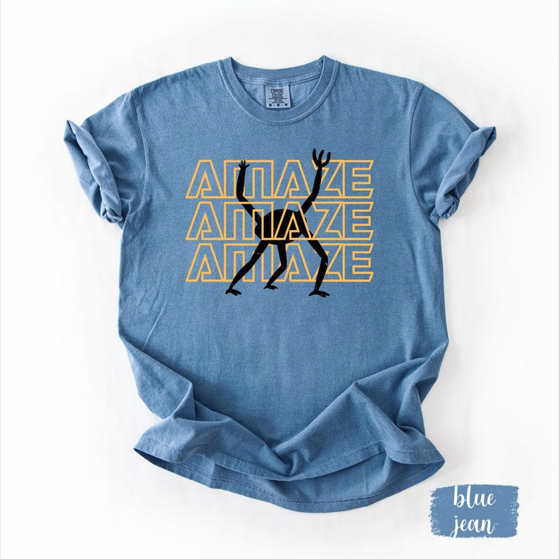May include: Blue jean-colored t-shirt with the word "AMAZE" stacked three times in a yellow font. A black stick figure is centered on the shirt, with its arms and legs outstretched. The shirt is made of a soft material.