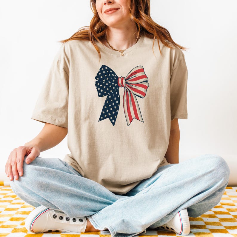 Peut inclure: Un t-shirt beige avec un n&oelig;ud rouge, blanc et bleu. Le n&oelig;ud est fait de tissu avec un motif de &eacute;toiles et de rayures.