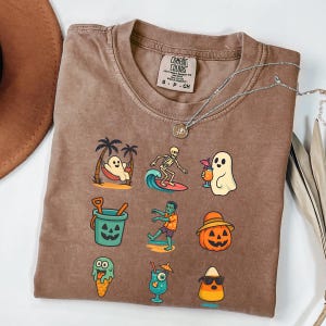 May include: Brown Comfort Colors t-shirt featuring a collection of Halloween-themed illustrations. The graphics include a ghost in a hammock, a skeleton surfing, a ghost with a cocktail, a jack-o'-lantern bucket, a zombie, and a candy corn with sunglasses.