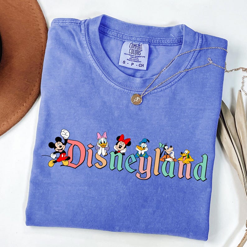 May include: A blue Comfort Colors t-shirt featuring the word "Disneyland" in colorful letters with cartoon characters Mickey Mouse, Daisy Duck, Minnie Mouse, Donald Duck, Goofy, and Pluto. A gold necklace is draped across the shirt.