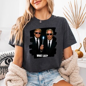 K&ouml;nnte beinhalten: Dunkelgraues T-Shirt mit einer Grafik von zwei M&auml;nnern in schwarzen Anz&uuml;gen und Sonnenbrillen, die einen leuchtenden Zauberstab halten. Der Text "WHAT LIST?" ist darunter gedruckt. Das T-Shirt ist l&auml;ssig.