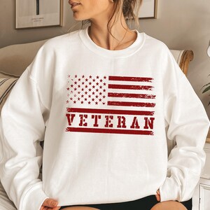 May include: White sweatshirt with a red, white, and blue American flag graphic and the word "VETERAN" in red.