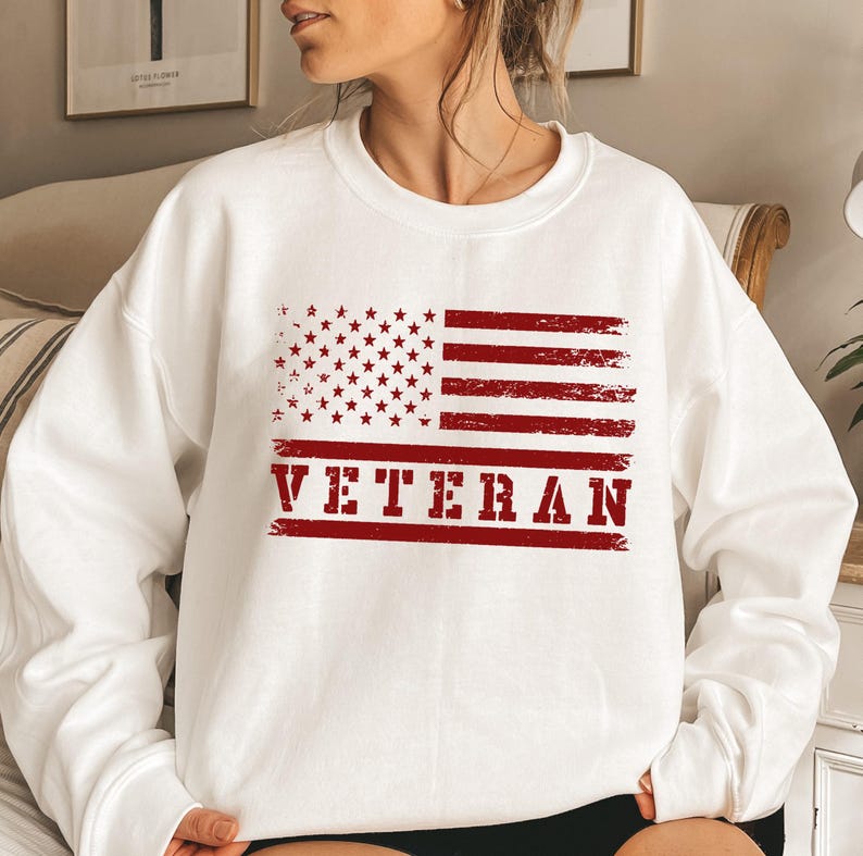 May include: White sweatshirt with a red, white, and blue American flag graphic and the word "VETERAN" in red.